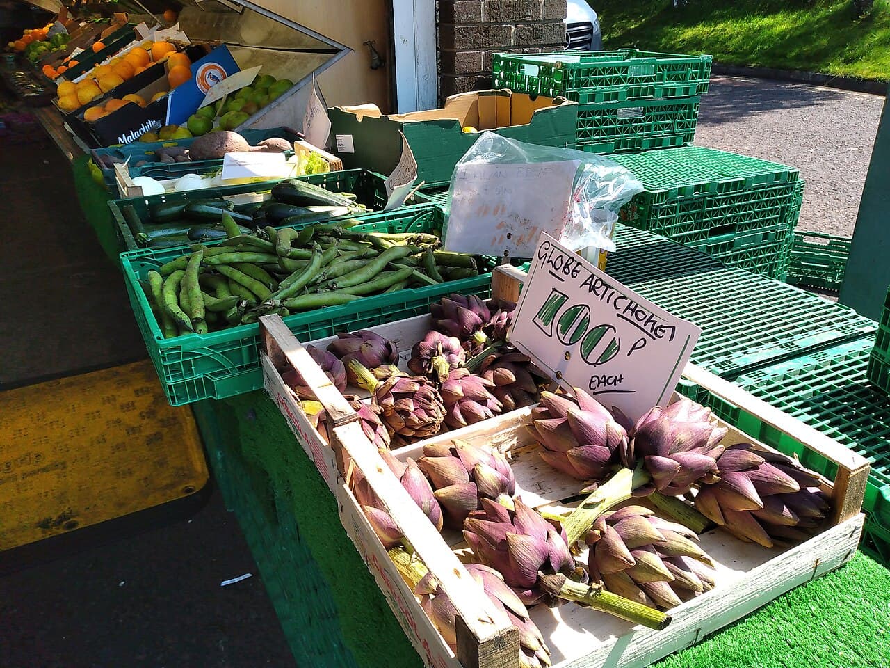 An outdoor market stall with wooden crates of purple-green globe artichokes and a handwritten sign, beside green crates of fava-bean pods